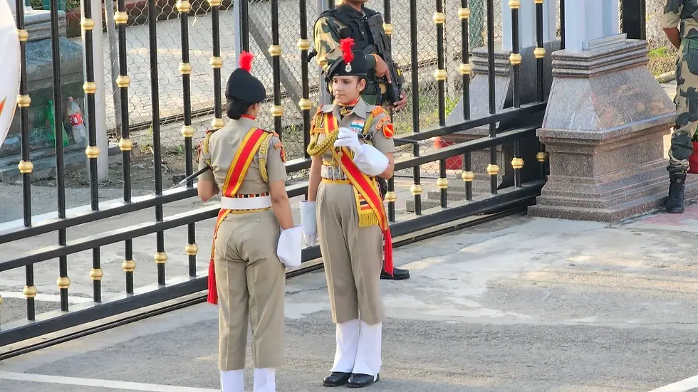 Two uniformed women perform a formal ceremony at a gated entrance. A guard stands nearby. The scene is structured and disciplined.