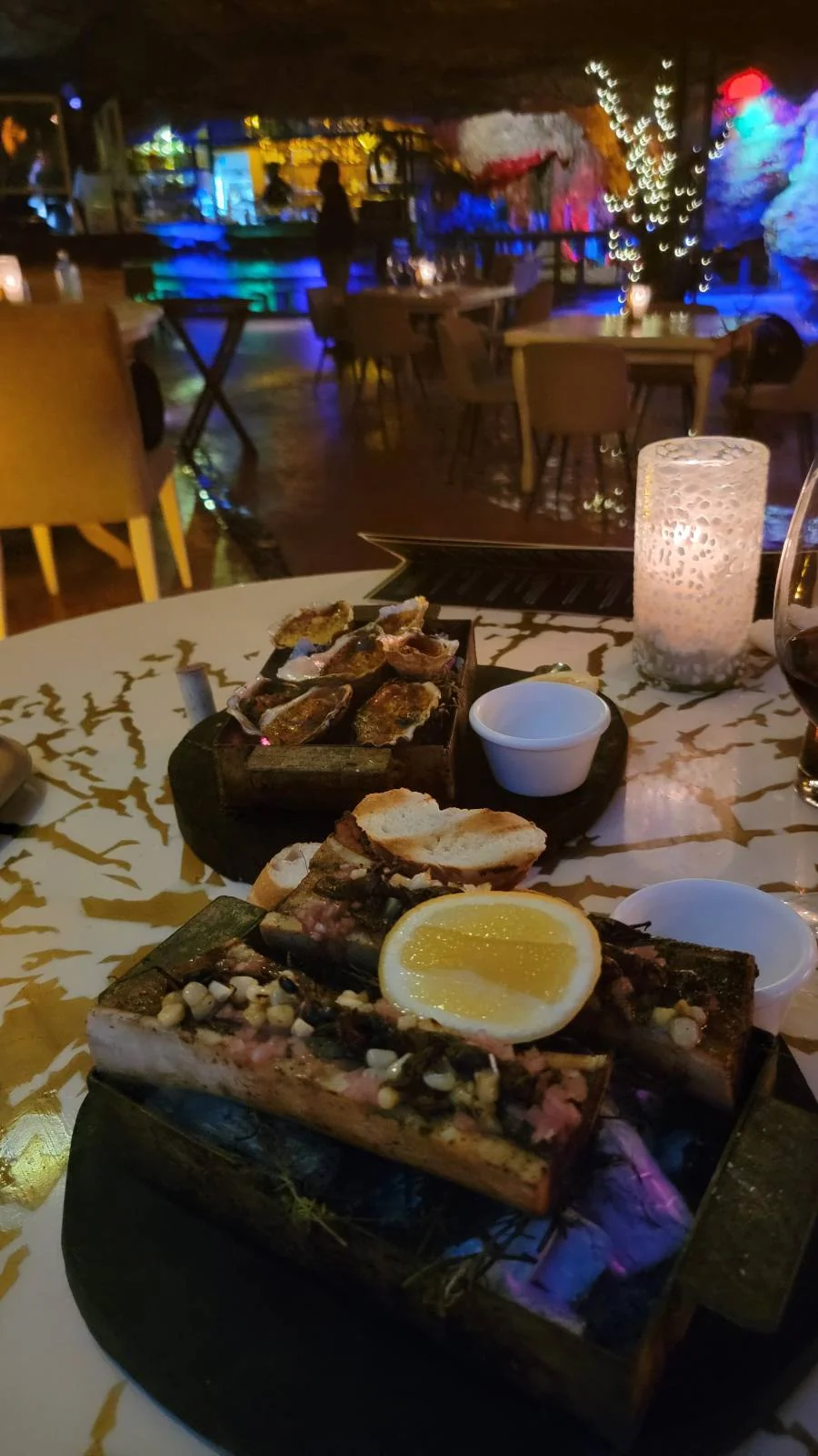 A dining table in a dimly lit restaurant is set with dishes of food, including bone marrow served in wooden trays, garnished with herbs and a lemon slice. Candles and bokeh lights create a cozy ambiance.