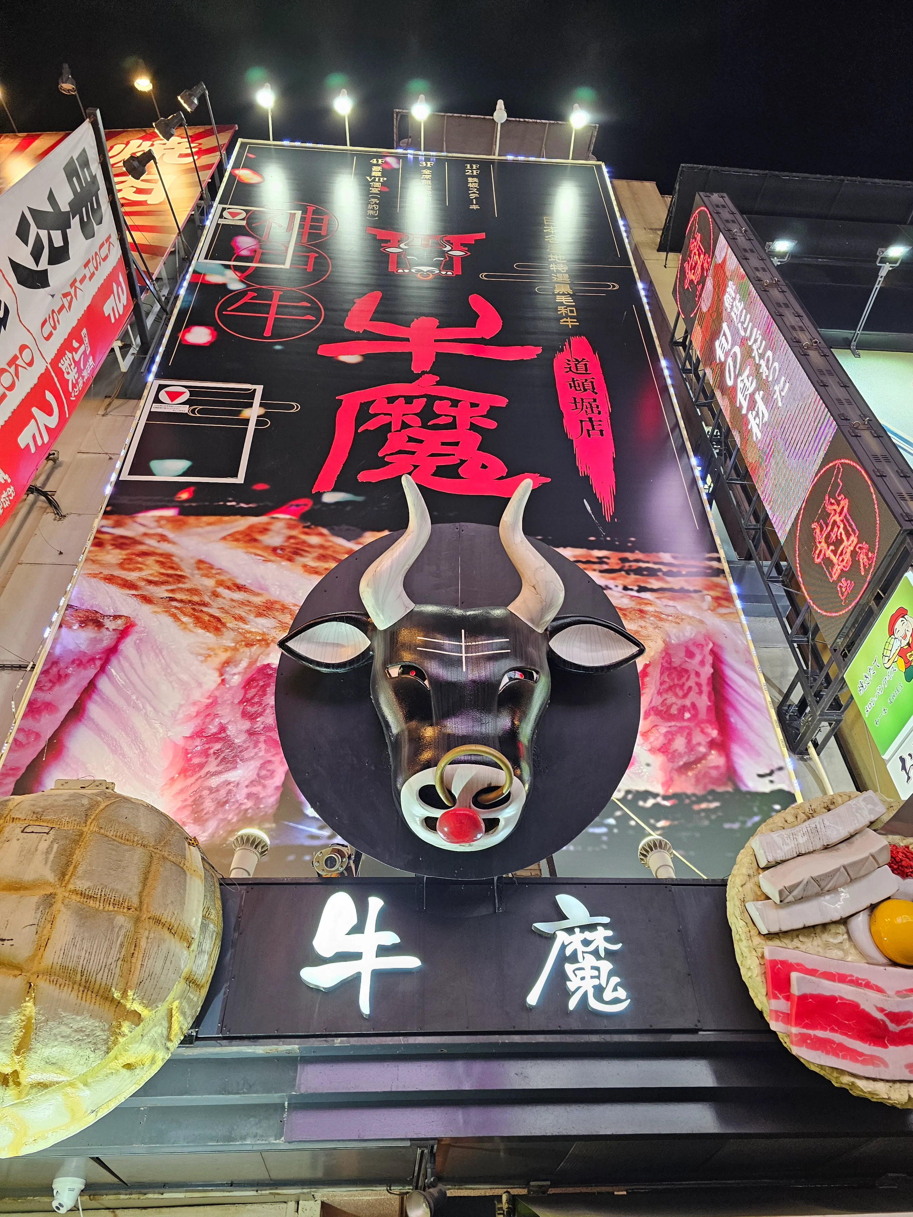 Brightly lit restaurant sign at night featuring a large 3D bull’s head, bold red Japanese characters, and images of food, with smaller illuminated signs and buildings surrounding it.