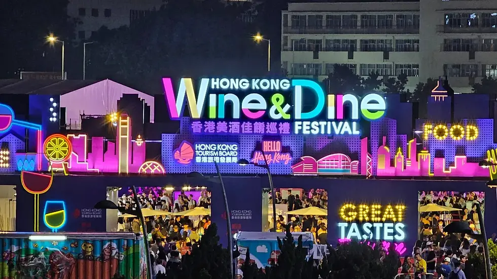 Neon-lit "Hong Kong Wine & Dine Festival" sign with lively crowd below. Bright, colorful skyline backdrop and festive atmosphere.