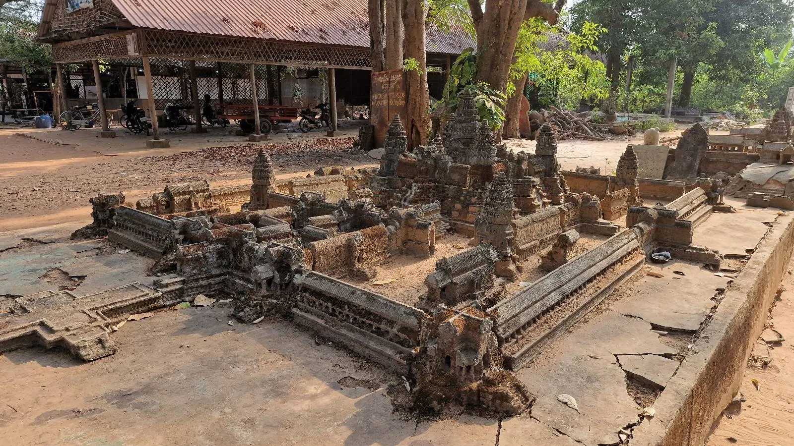 A detailed miniature replica of an ancient temple complex is displayed outdoors. The structure showcases intricate carvings and architecture. Trees and a rustic building are visible in the background under a clear sky.