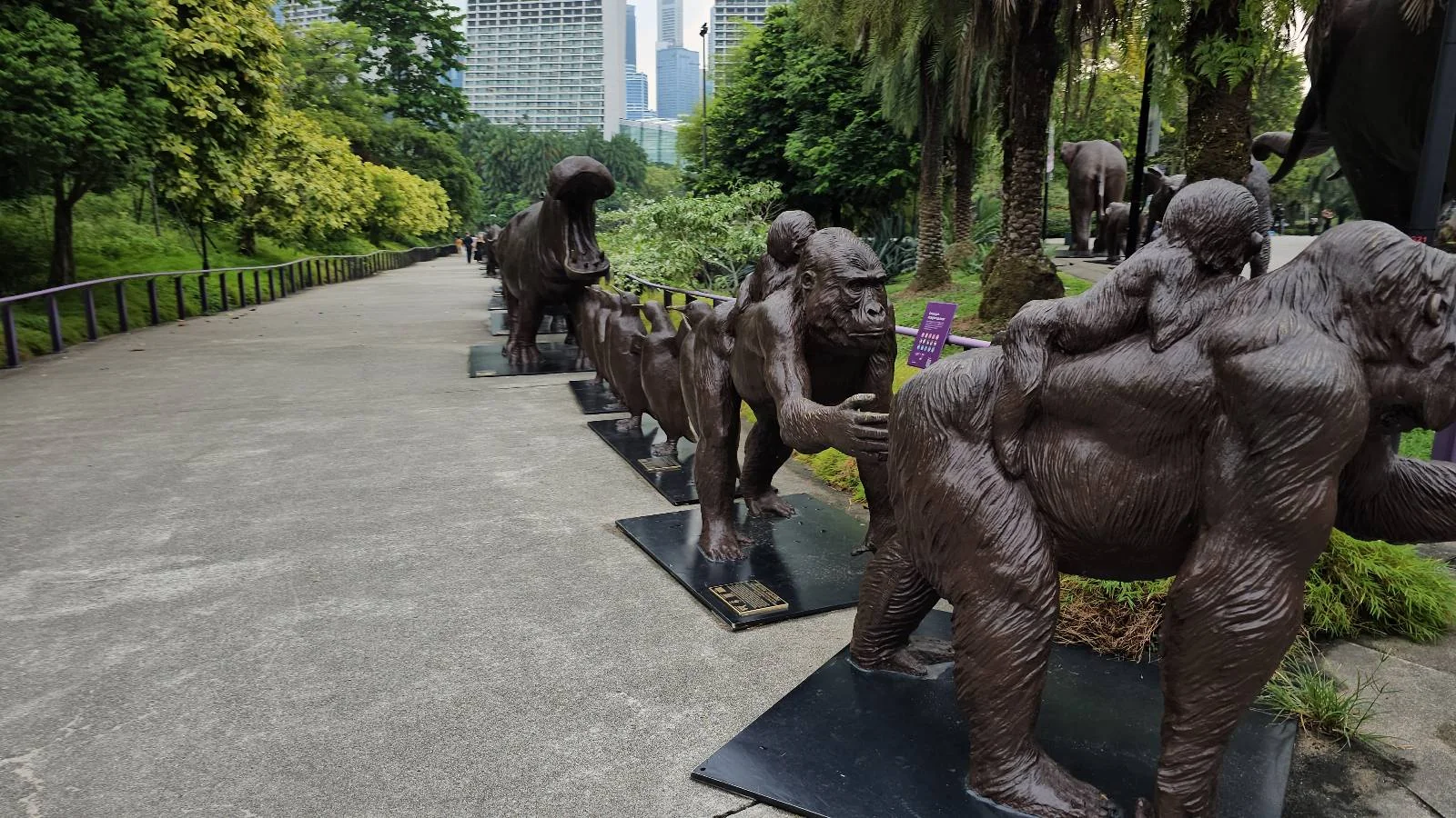 A series of bronze gorilla sculptures arranged along a park pathway, depicting various poses and sizes. Trees and city buildings are visible in the background, providing a contrast between nature and urban environment.