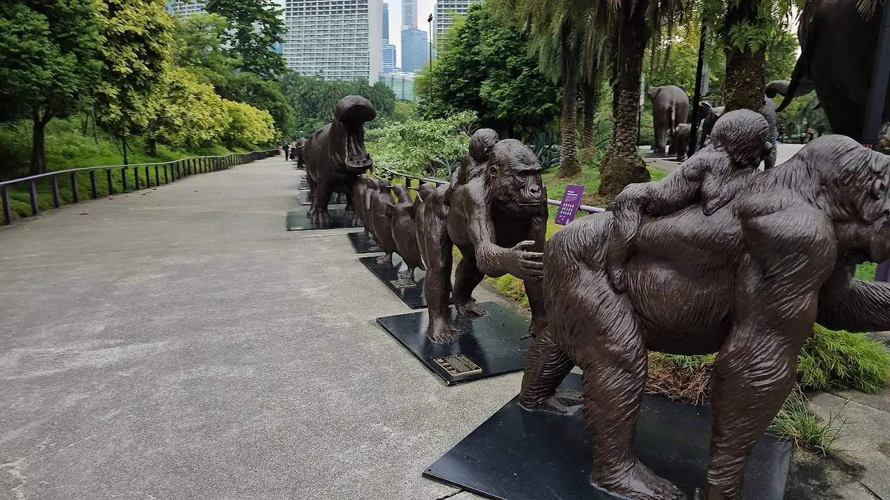 A series of bronze gorilla sculptures arranged along a park pathway, depicting various poses and sizes. Trees and city buildings are visible in the background, providing a contrast between nature and urban environment.