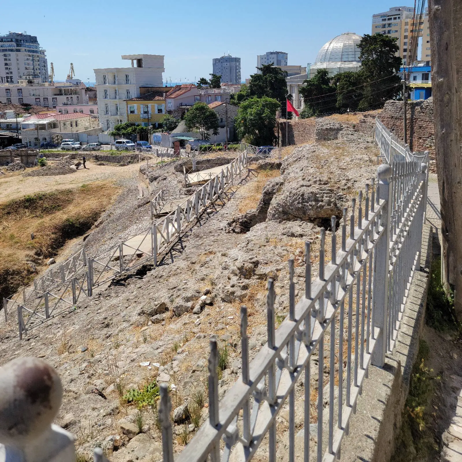 Ancient ruins with a walkway and metal railing, set against a cityscape background of modern buildings and a dome, under a clear blue sky.