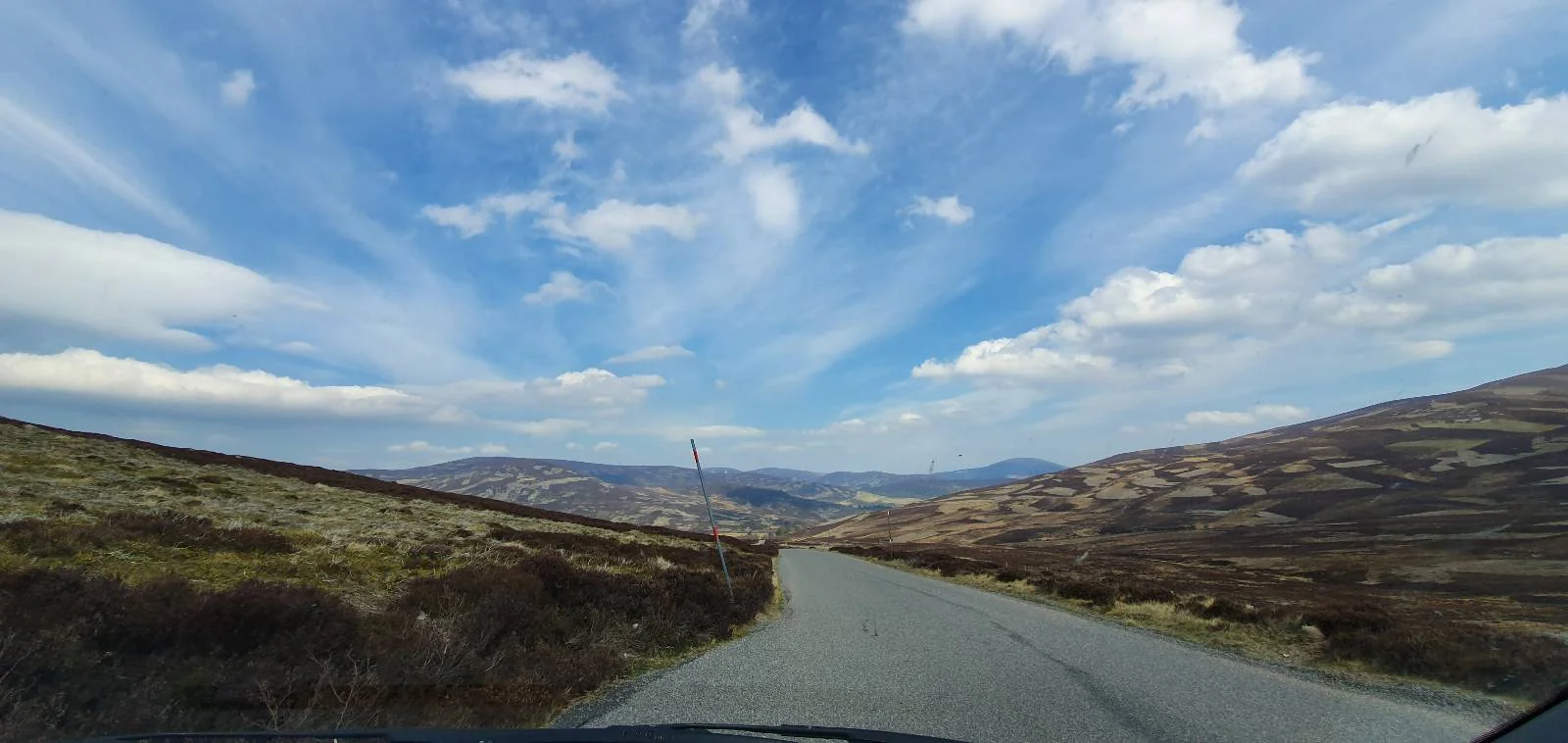 A winding road stretches through a hilly landscape under a dramatic sky with scattered clouds and sun rays. The scene is surrounded by grassy hills, creating a sense of openness and tranquility.
