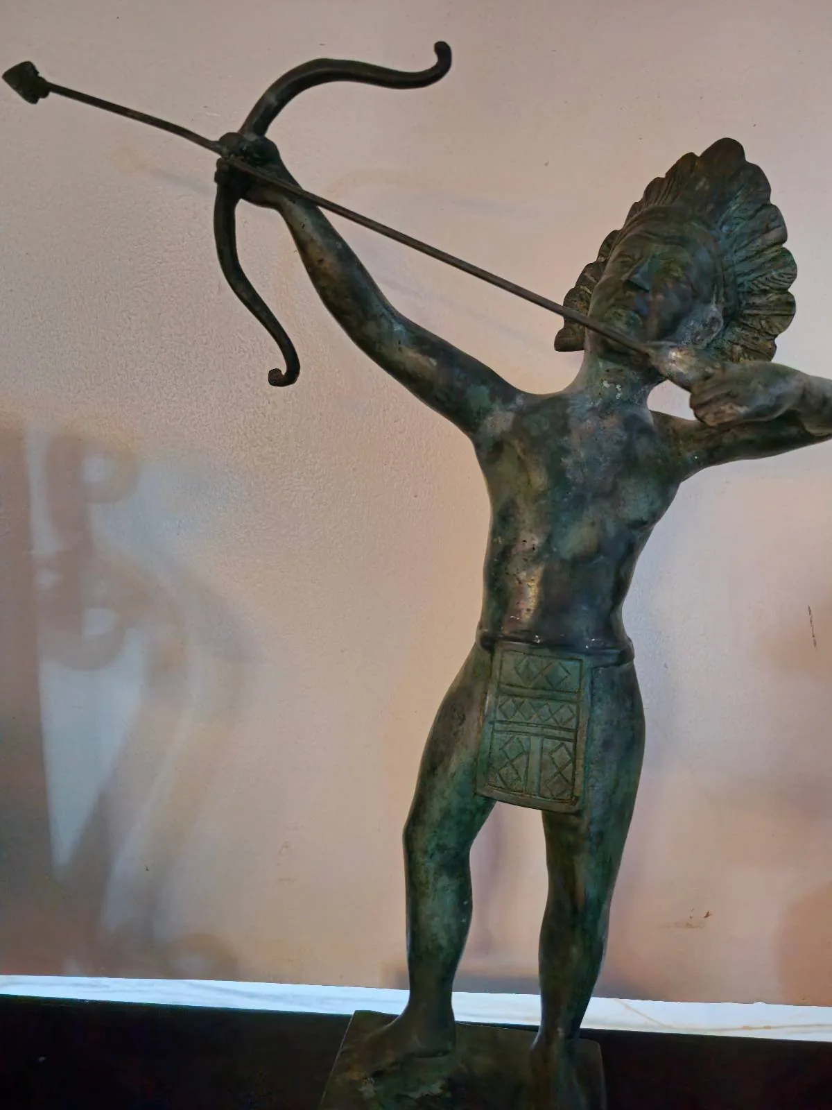 A bronze statue of a standing archer figure wearing a headdress and a rectangular loincloth, pulling back a bow with an arrow aimed forward. The background is plain, enhancing the sculpture's detail.
