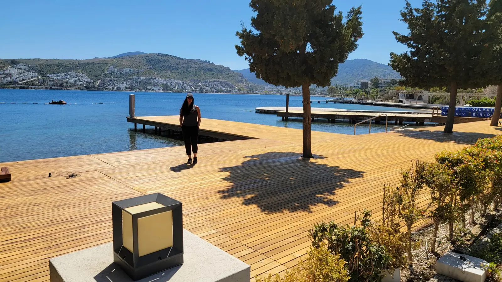 A person stands on a wooden dock by a calm blue lake, surrounded by trees and hills under a clear sky.
