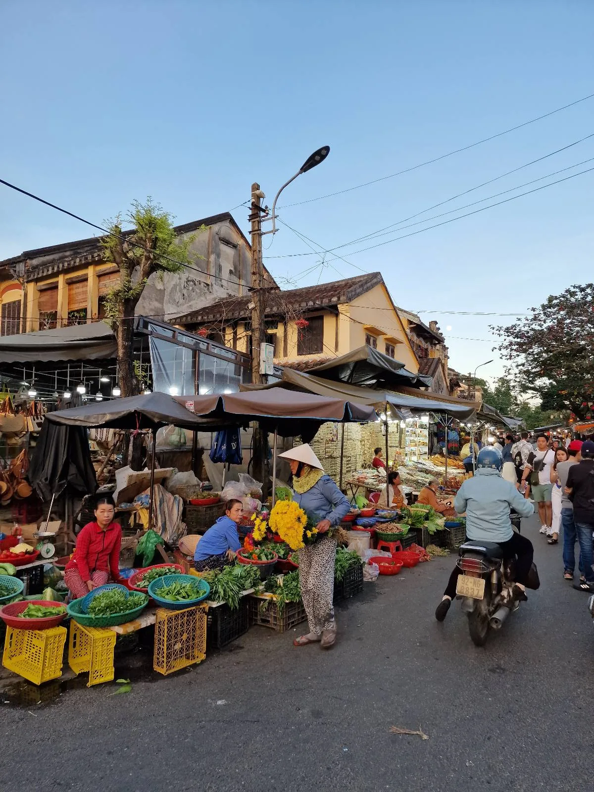 Street market scene with vendors selling vegetables in baskets. People are browsing and buying produce. A motorbike and pedestrians are visible, with traditional buildings and a clear sky in the background.