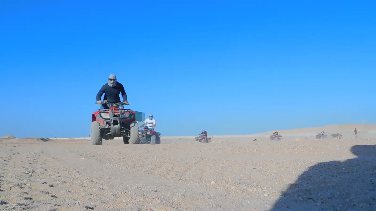 Two people riding red ATVs in a desert landscape. Both are wearing helmets and protective gear. The terrain is sandy with rocky hills in the background under a clear blue sky.