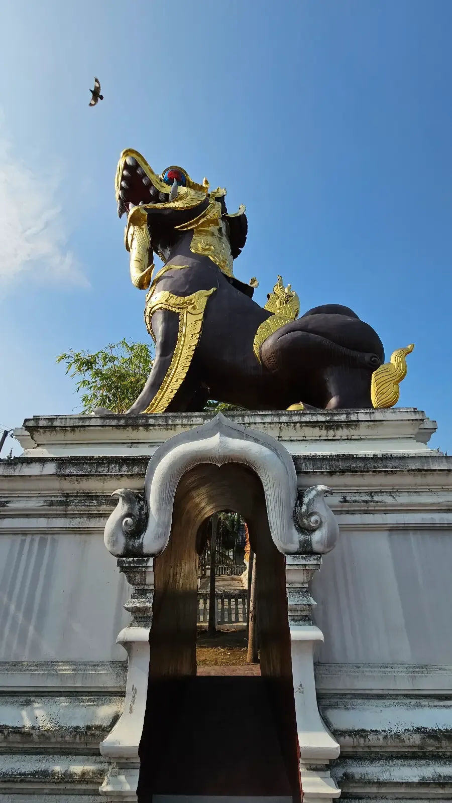 A large black and gold mythical lion statue sits on a stone pedestal with an arched doorway, against a blue sky. The statue has ornate details and looks upward.