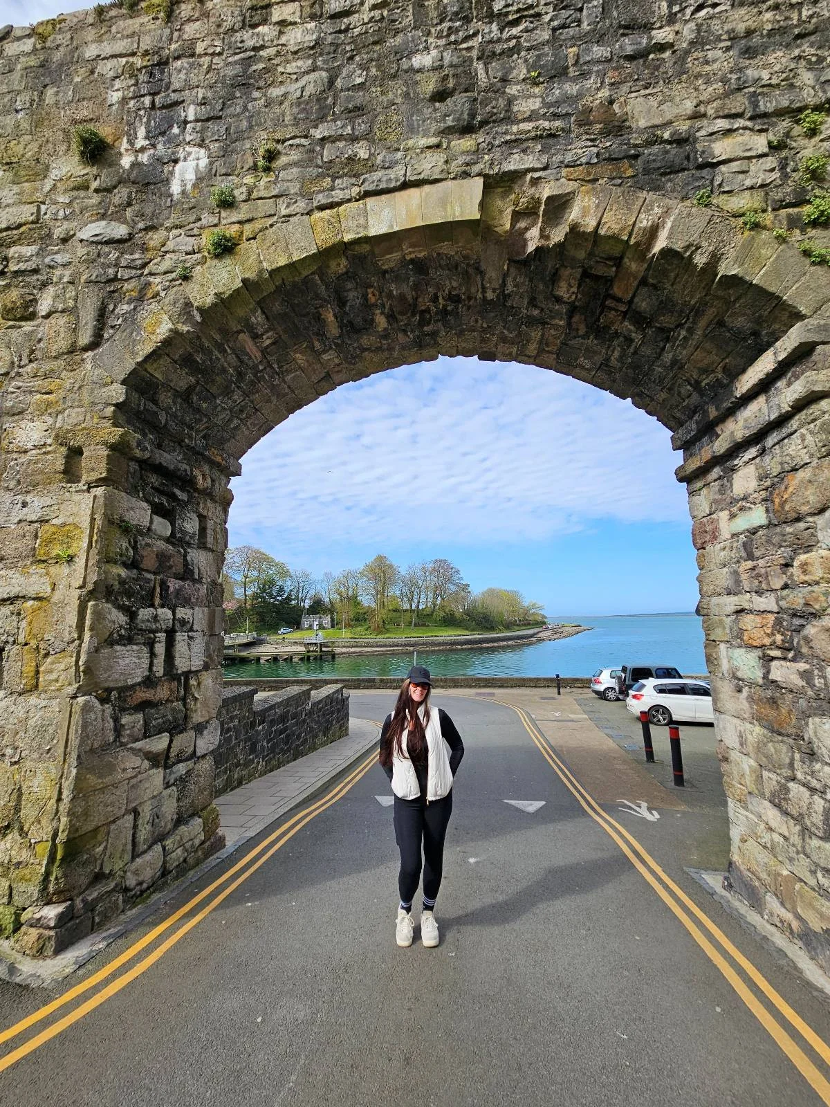 A person stands under a large stone archway beside a road with yellow lines. In the background, there are trees, a body of water, and a partly cloudy sky.