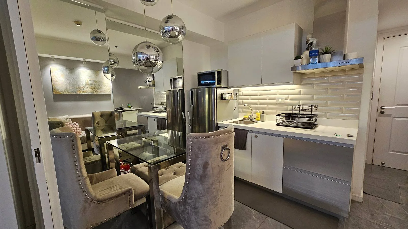 Modern kitchen and dining area with glass table, tufted chairs, mirrored backsplash, and pendant lights. Bright, clean, and stylish decor.