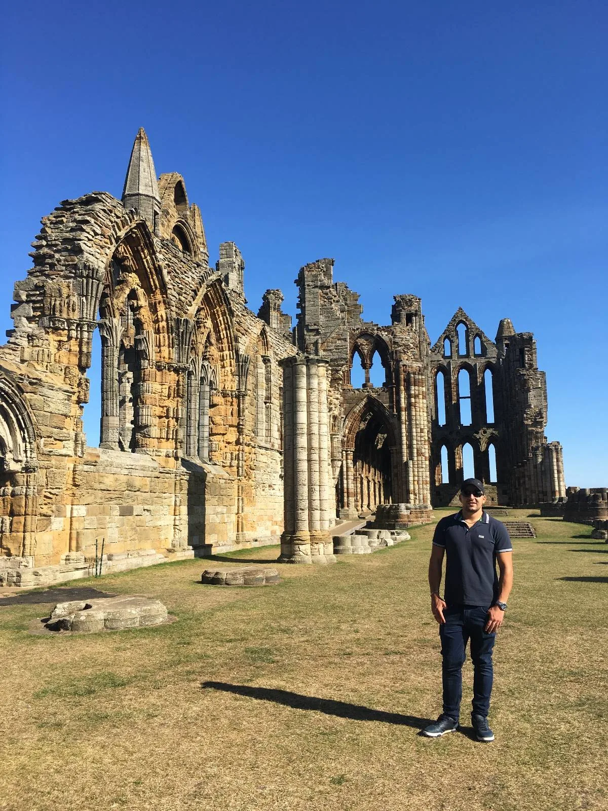 A person stands in front of the ruins of an ancient stone abbey under a clear blue sky. The abbey features large, arched windows and weathered stone walls. The ground is grassy and the scene is sunlit.