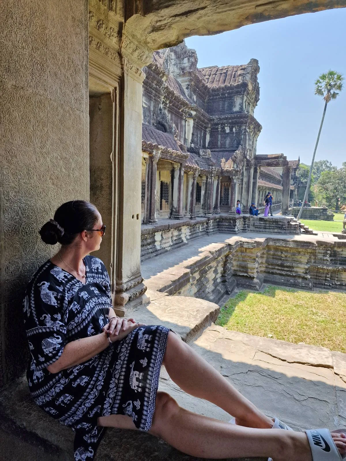 A woman in a black and white dress sits on a stone window ledge, looking out at the ancient ruins of Angkor Wat under a clear blue sky.