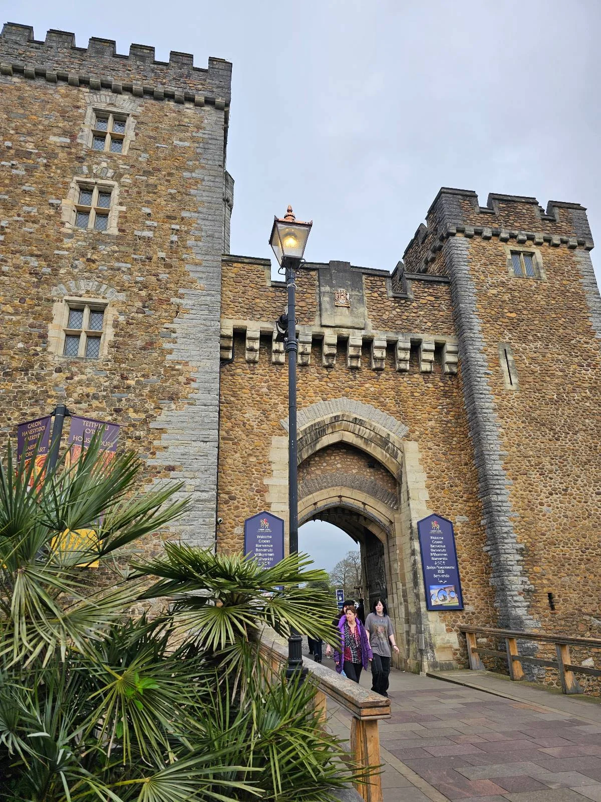 Lamp post and people walking towards the entrance of an ancient stone castle with banners.