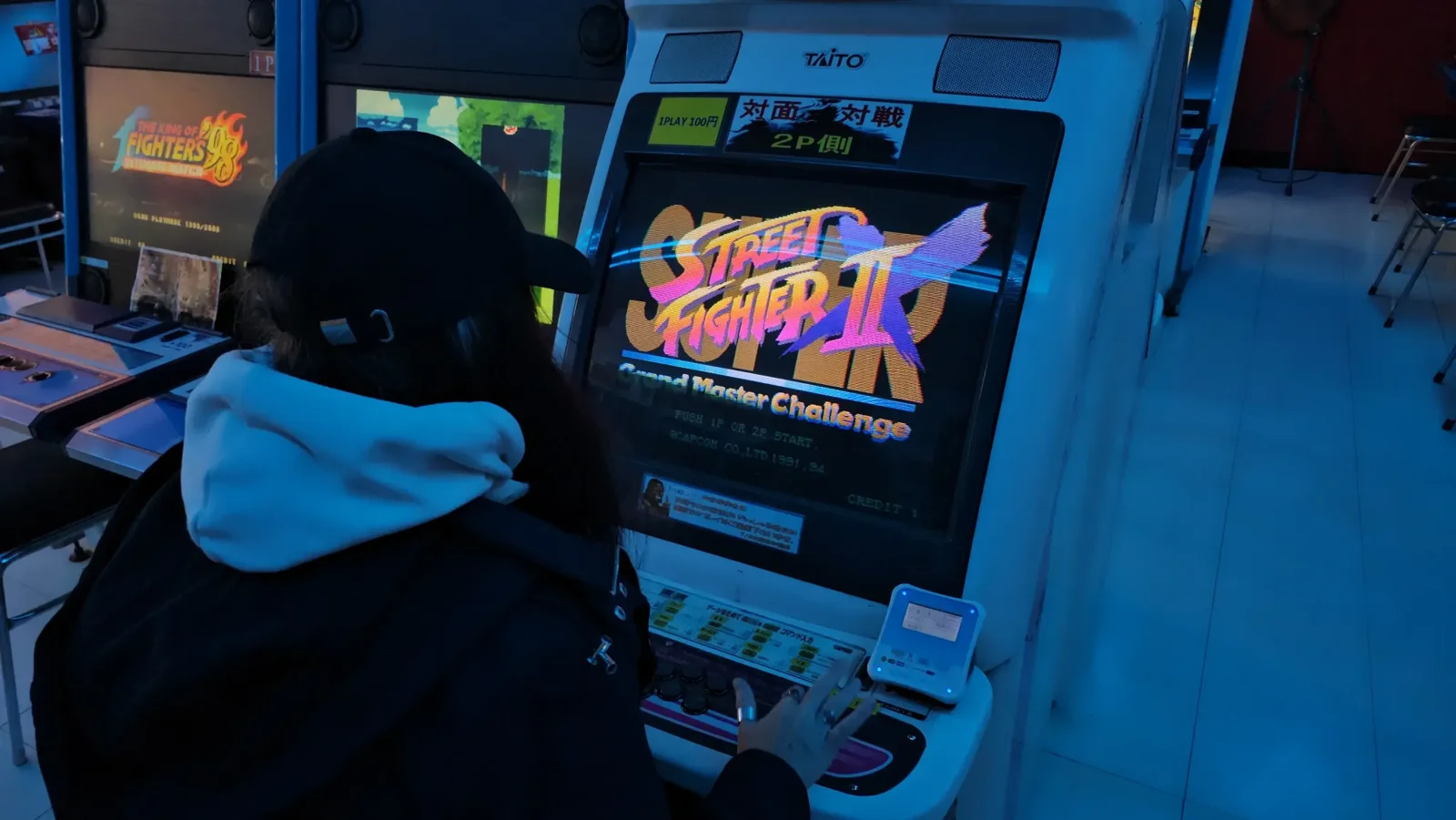A person in a hoodie and cap plays Street Fighter II on an arcade machine in a dimly lit gaming area.