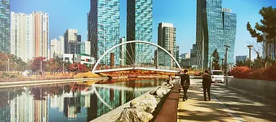 A futuristic urban park scene in Songdo showing a pedestrian arch bridge over a calm canal, flanked by modern glass skyscrapers.
