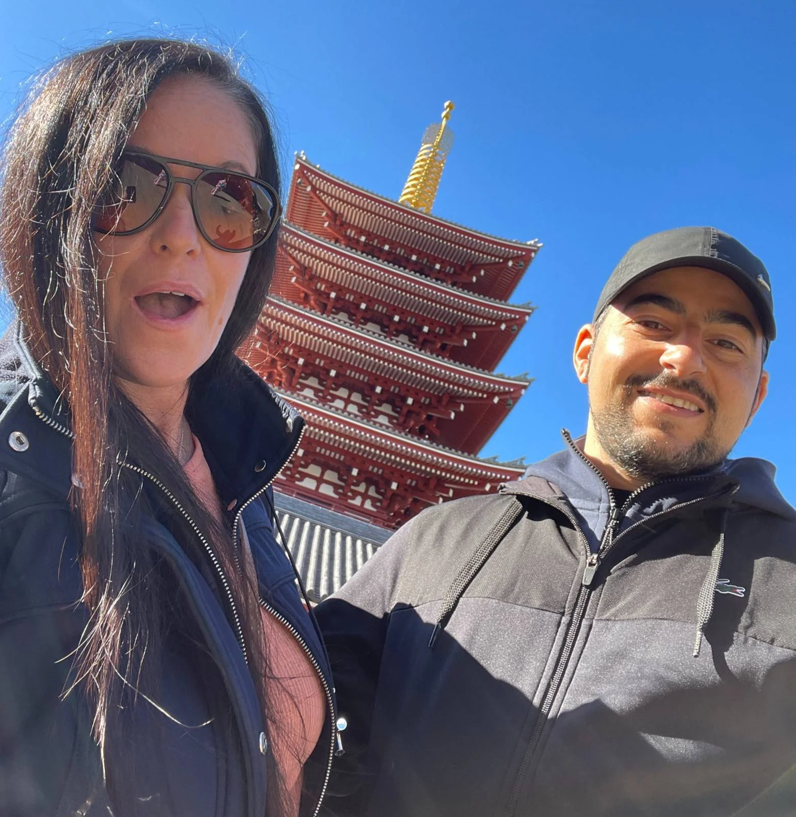 A man and woman in sunglasses smile in front of a red pagoda under a clear blue sky. The backdrop is vibrant and sunny.