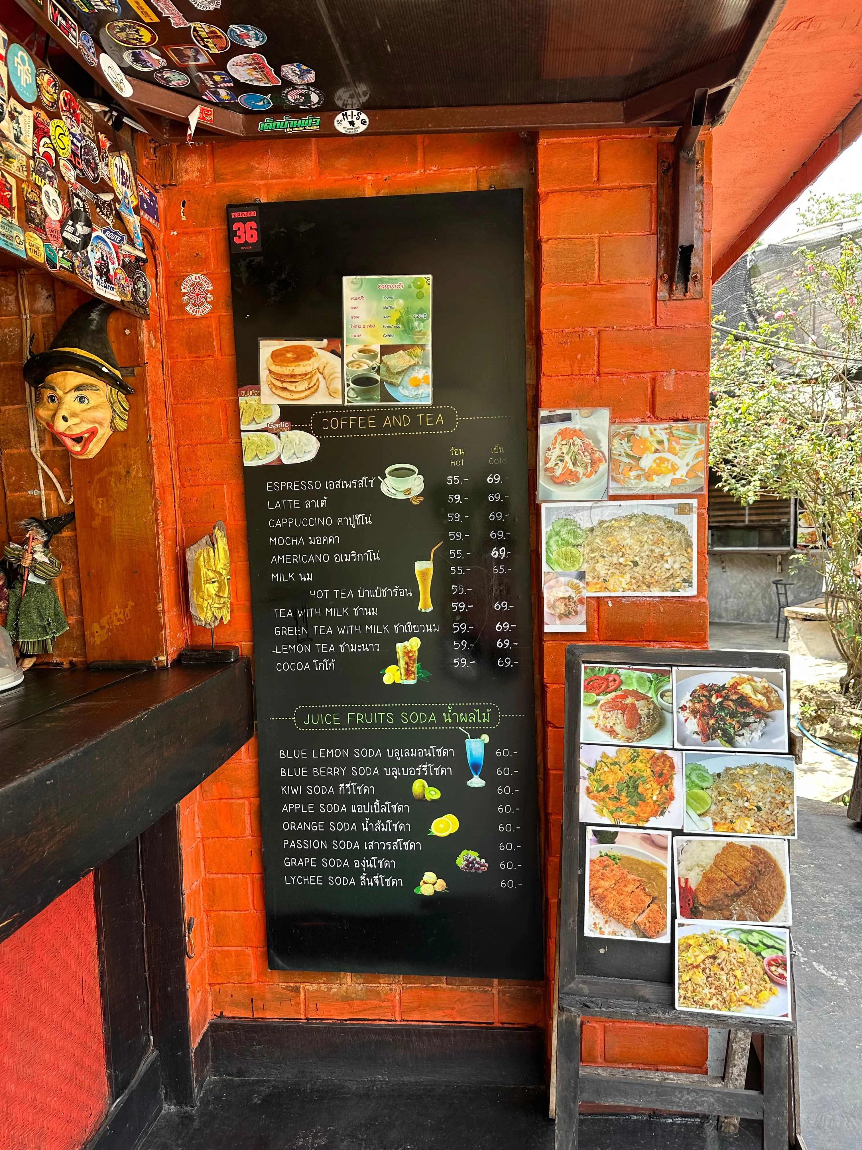 A black menu board with food items and prices is mounted on an orange restaurant wall. Colorful food photos and cartoon decorations surround the menu, which is next to a small counter and an outdoor seating area.