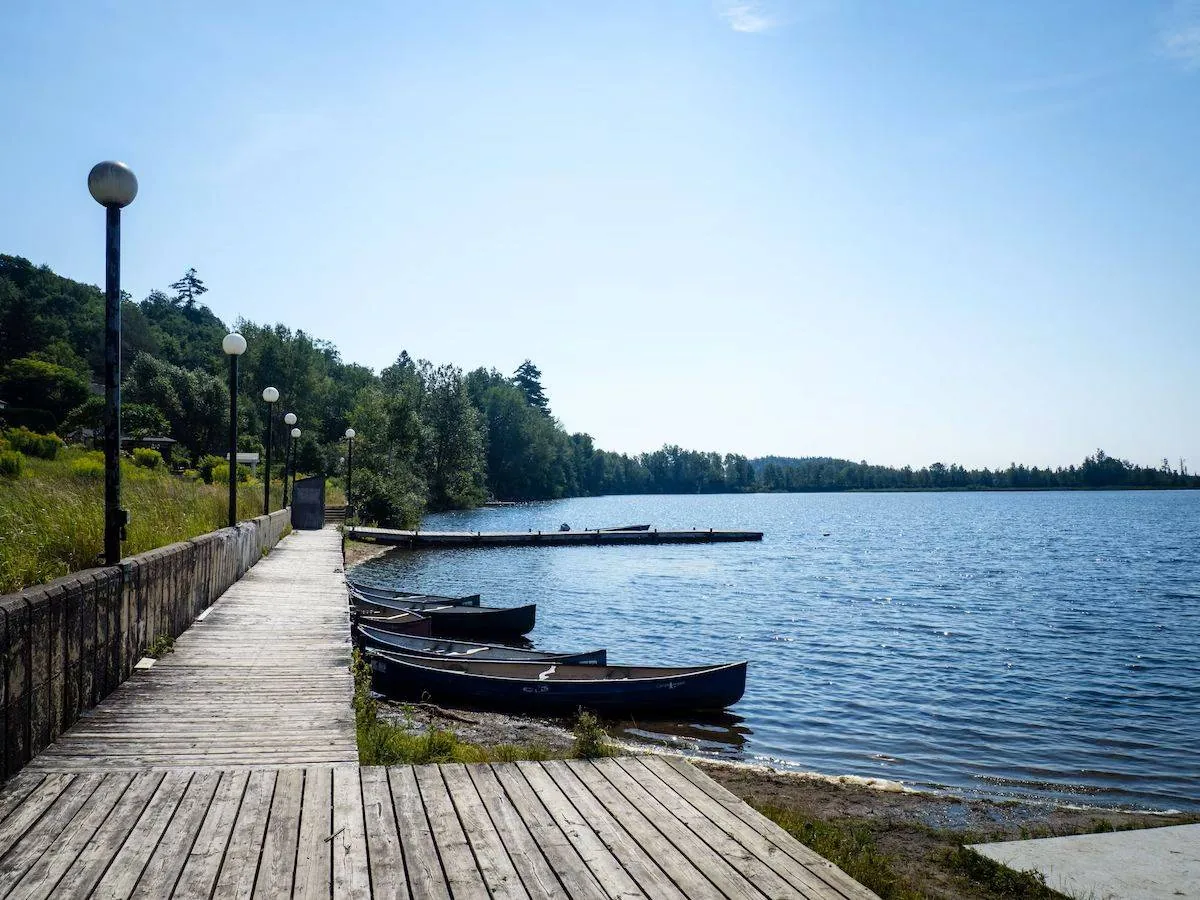 Wooden boardwalk along a lake with canoes, lamp posts, and lush greenery. Clear blue sky creates a tranquil atmosphere.