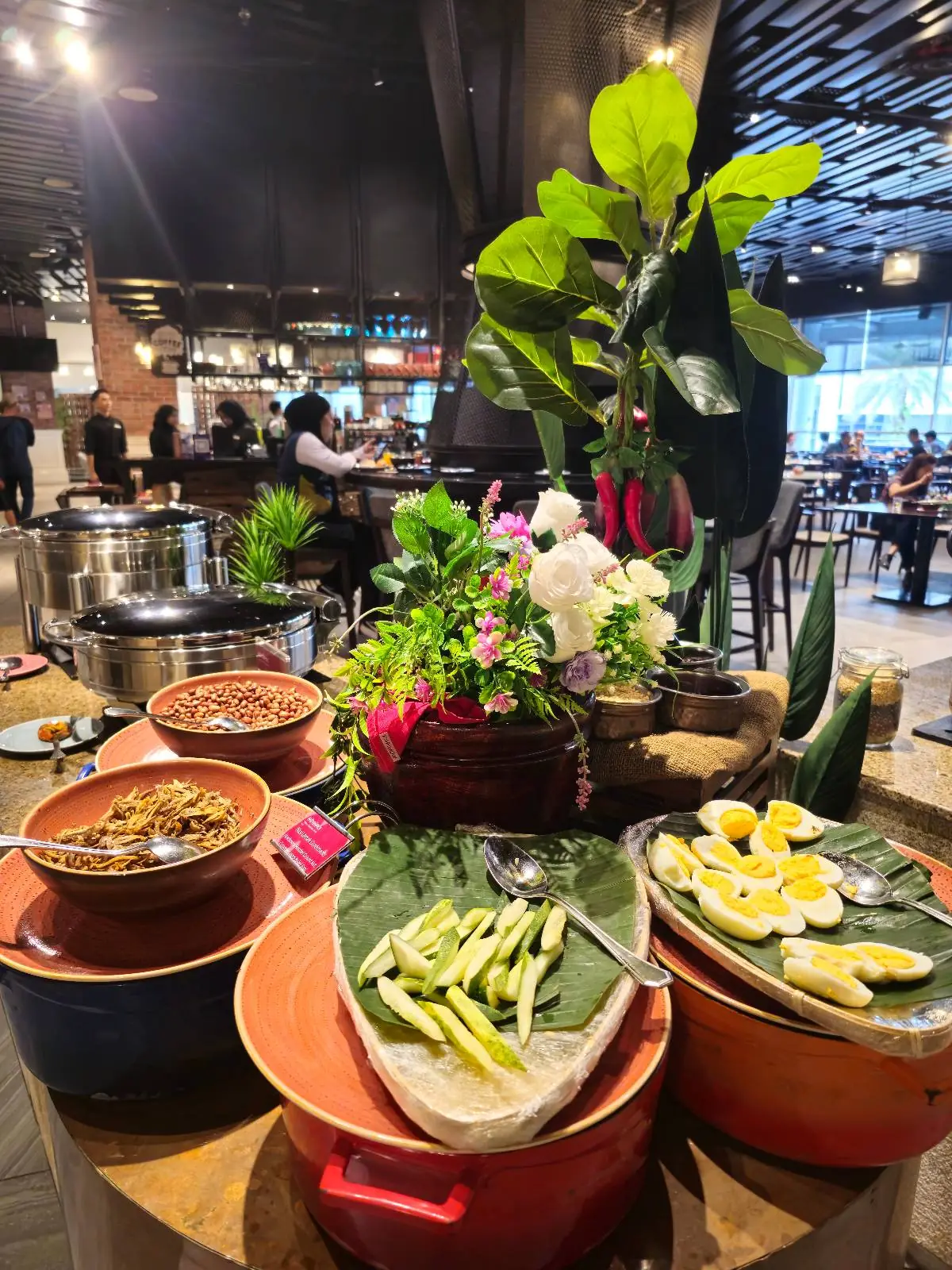 A vibrant buffet setup features a variety of dishes in clay pots and on banana leaves, with fresh green herbs and bright yellow flowers adding color. The background shows a bustling restaurant atmosphere with people and modern decor.