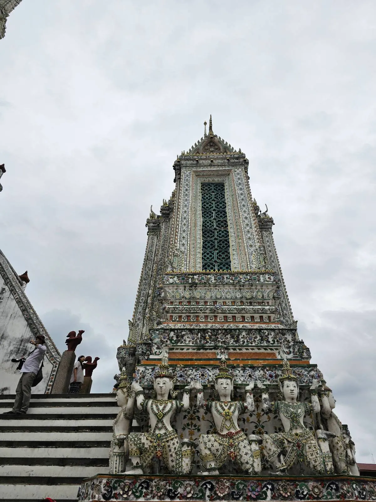 Tall temple structure with intricate carvings and colorful decorations, viewed from a low angle. A person is walking up the stairs on the left side. Cloudy sky in the background.