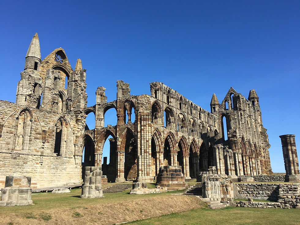 Ruins of a stone abbey with arched windows against a clear blue sky. Sunlit grass surrounds the ancient structure, creating a serene mood.