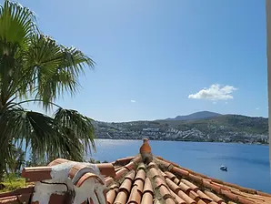 A sunny view of terracotta roof tiles in the foreground, with a palm tree on the left and the calm blue sea, coastal town, and hills of Gundogan, Bodrum in the background under a clear blue sky, radiating Turkish charm.