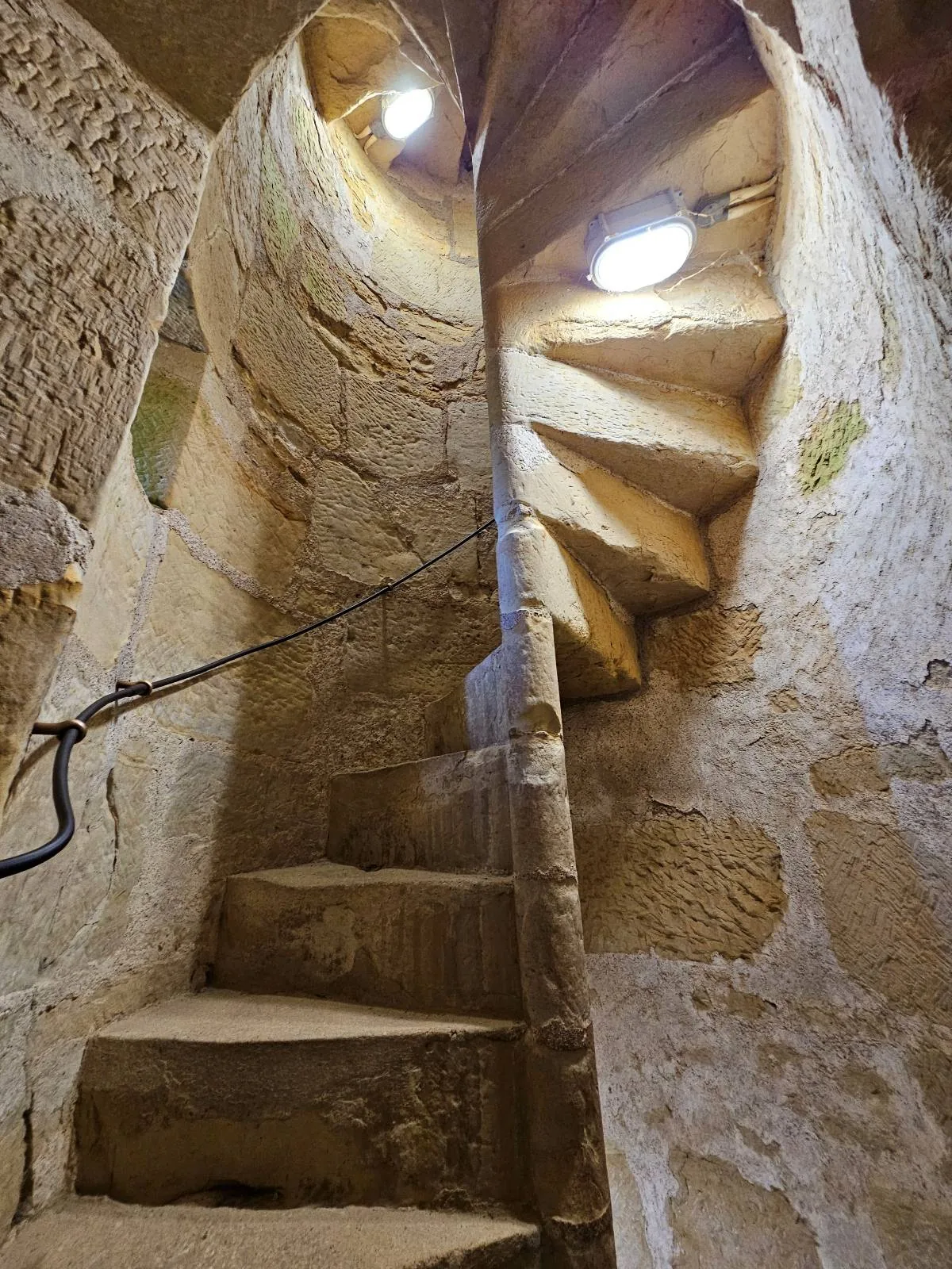 Spiral stone staircase inside an old, narrow tower with rough, textured walls and small circular windows letting in natural light.