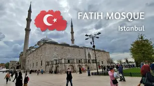 Fatih Mosque in Istanbul under cloudy sky, with visitors walking; white text and Turkish flag graphic overlay.