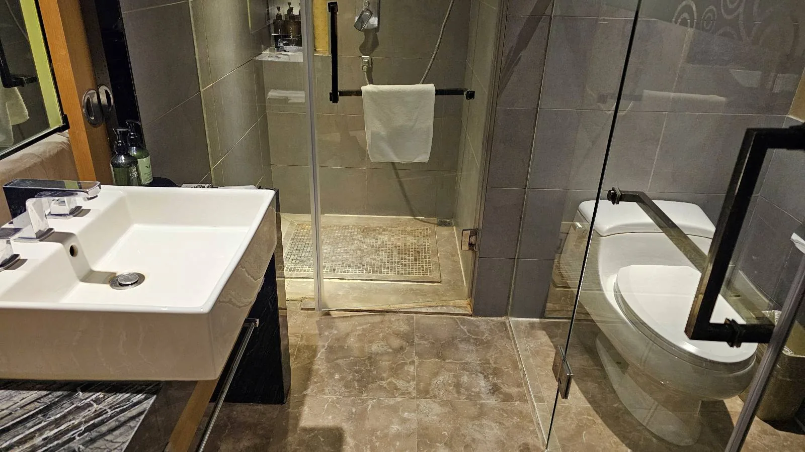 Modern bathroom with a glass shower enclosure, white sink on a dark vanity, wall-mounted toilet, and a beige tiled floor. A towel hangs on the glass shower door.