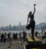 A statue holding a lighted orb stands in the foreground with a group of people gathered nearby. The background features a cityscape with tall buildings along a waterfront under a cloudy sky.