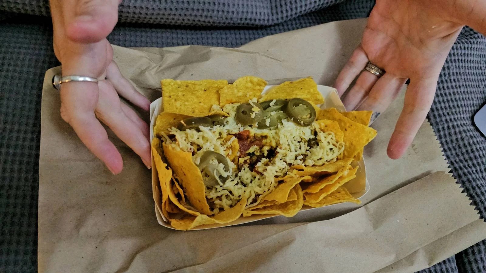 A tray of nachos topped with melted cheese and jalapeño slices is placed on a brown napkin, with two hands framing the tray from either side.