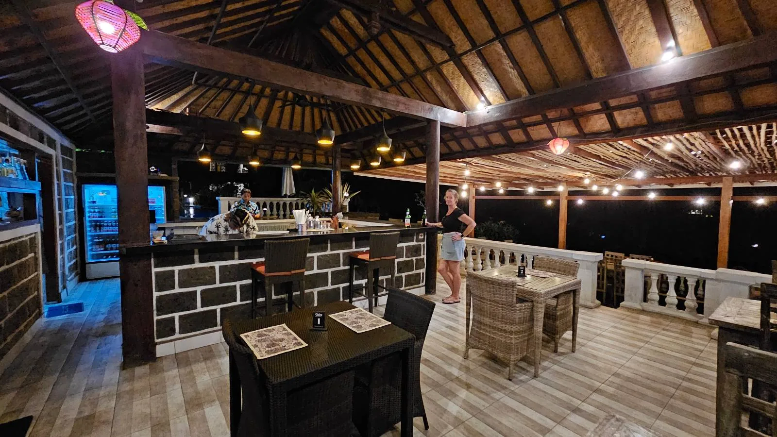 A cozy, open-air restaurant with wooden beams and string lights. Tables and chairs are set up under a high, thatched roof. A person stands by the bar counter, and a colorful lantern hangs in the foreground. The atmosphere is warm and inviting.