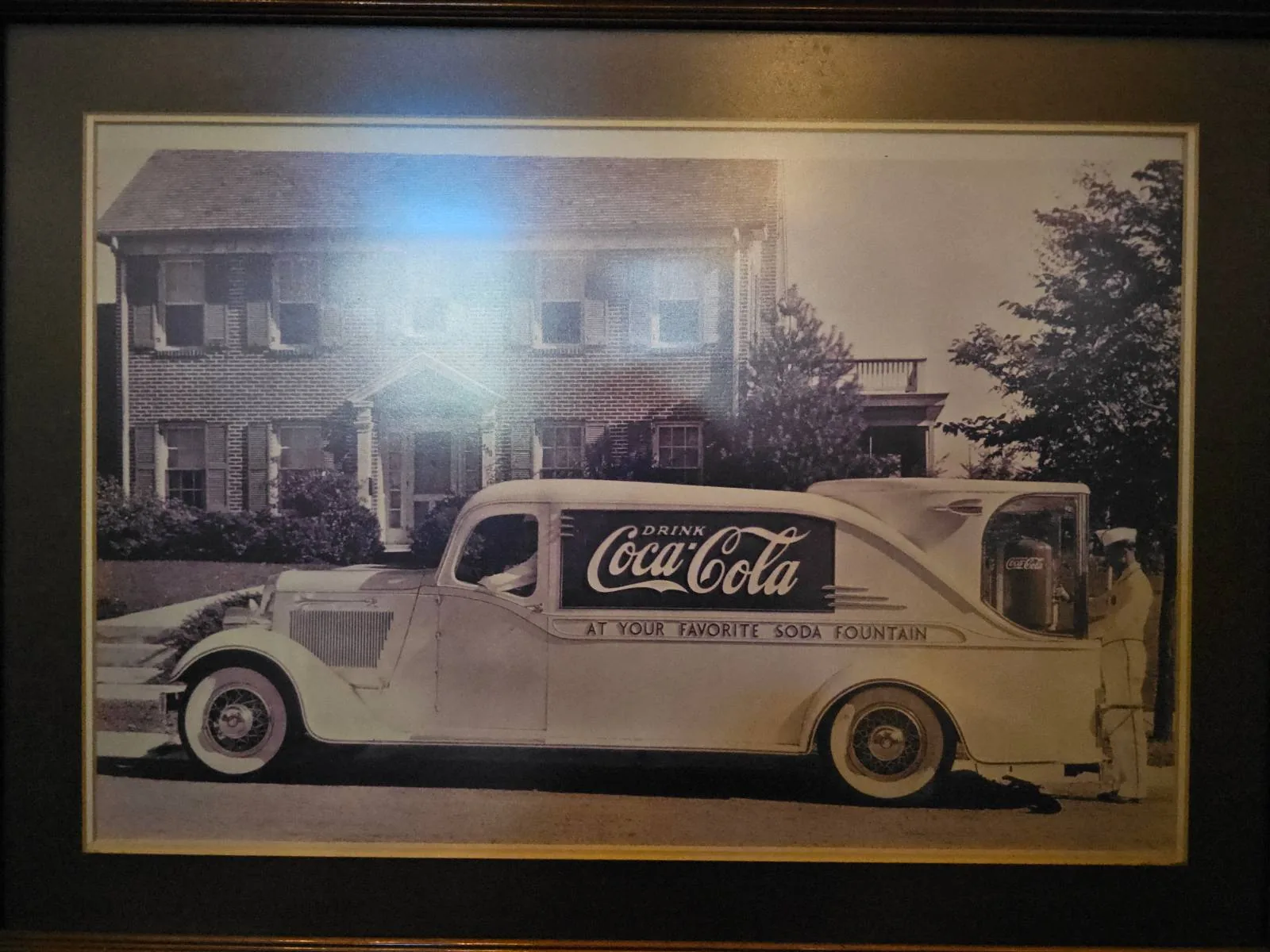 A vintage Coca-Cola delivery truck is parked in front of a two-story brick house. The truck has classic curved lines and displays the Coca-Cola logo prominently on its side. Trees and shrubbery are visible in the background.