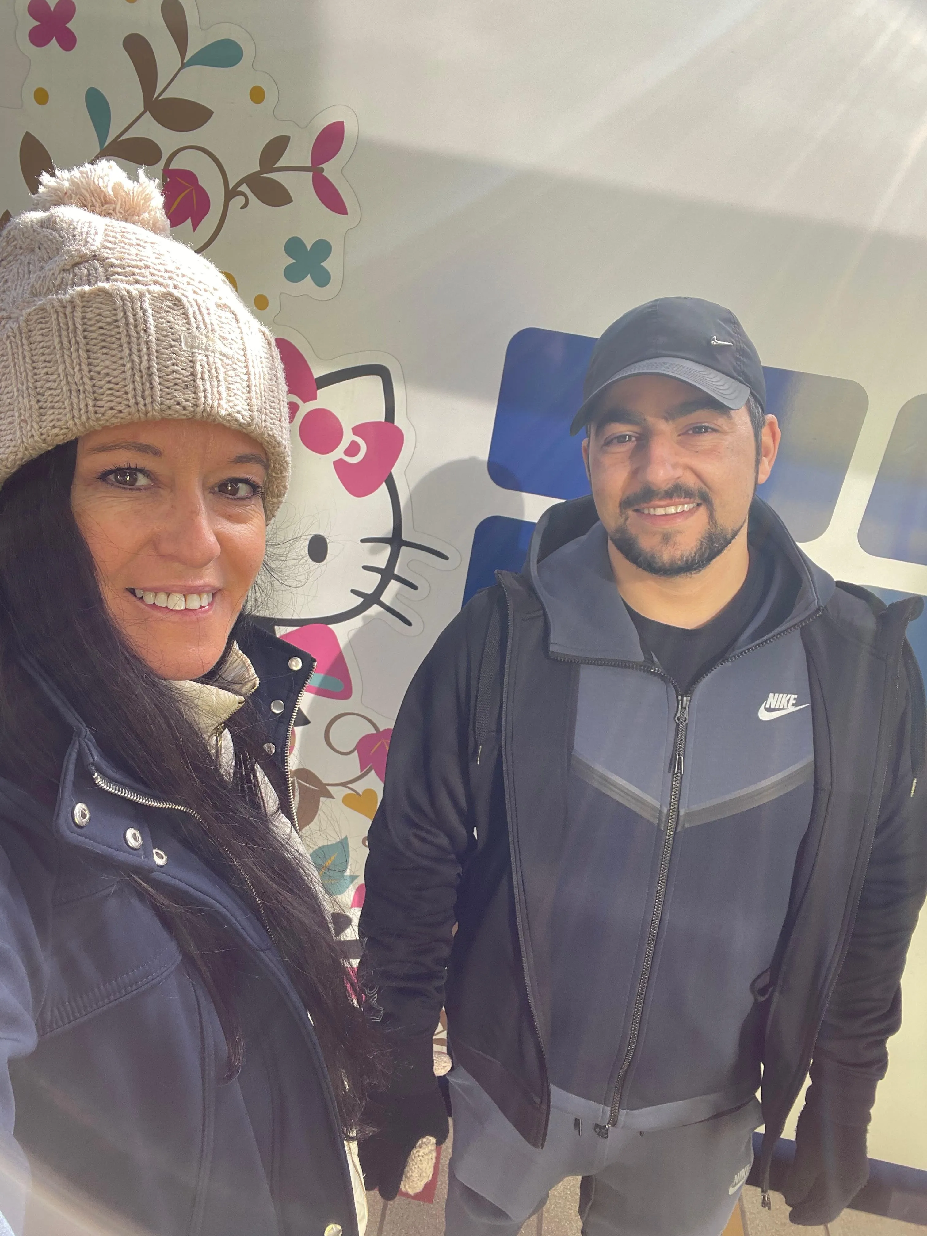 Two people smiling and posing for a selfie in front of a wall with a Hello Kitty design. The person on the left wears a beige knit hat, while the person on the right wears a black cap and jacket.