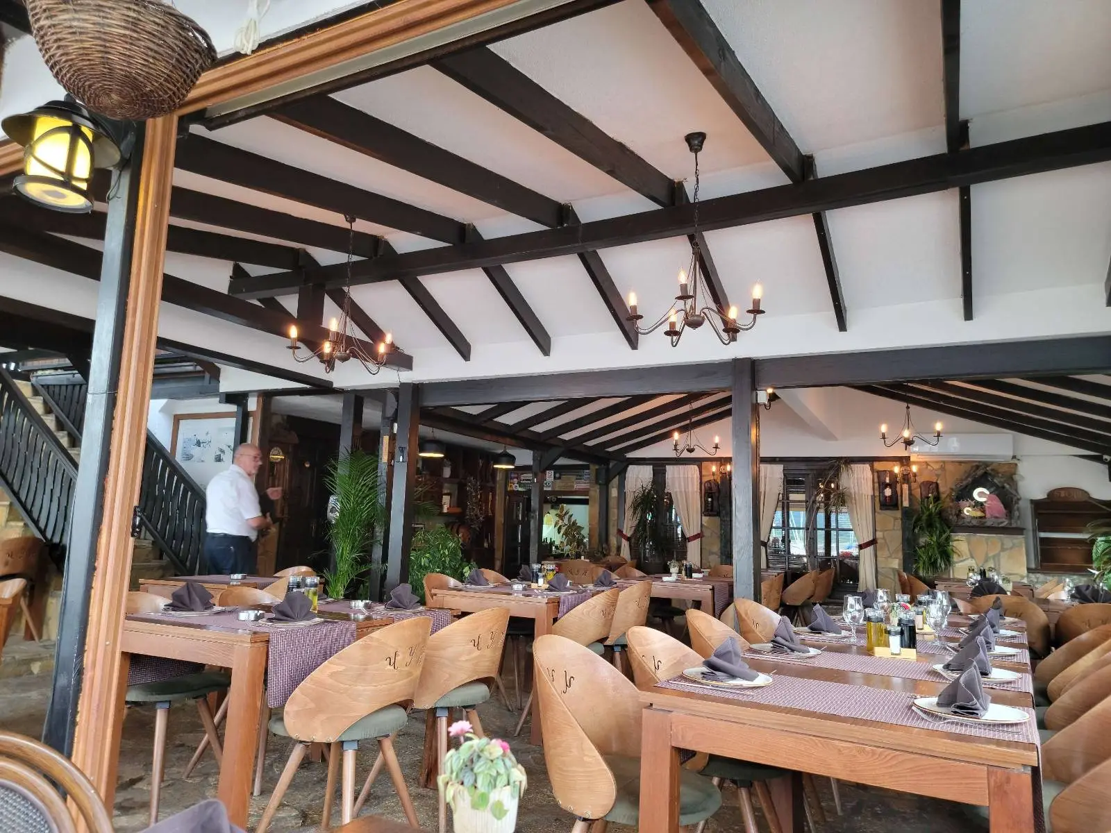 A cozy restaurant interior with wooden beams and chandeliers. Several wooden tables and chairs are set with tableware and napkins. A few potted plants and decorations are visible. A person stands near a staircase in the background.