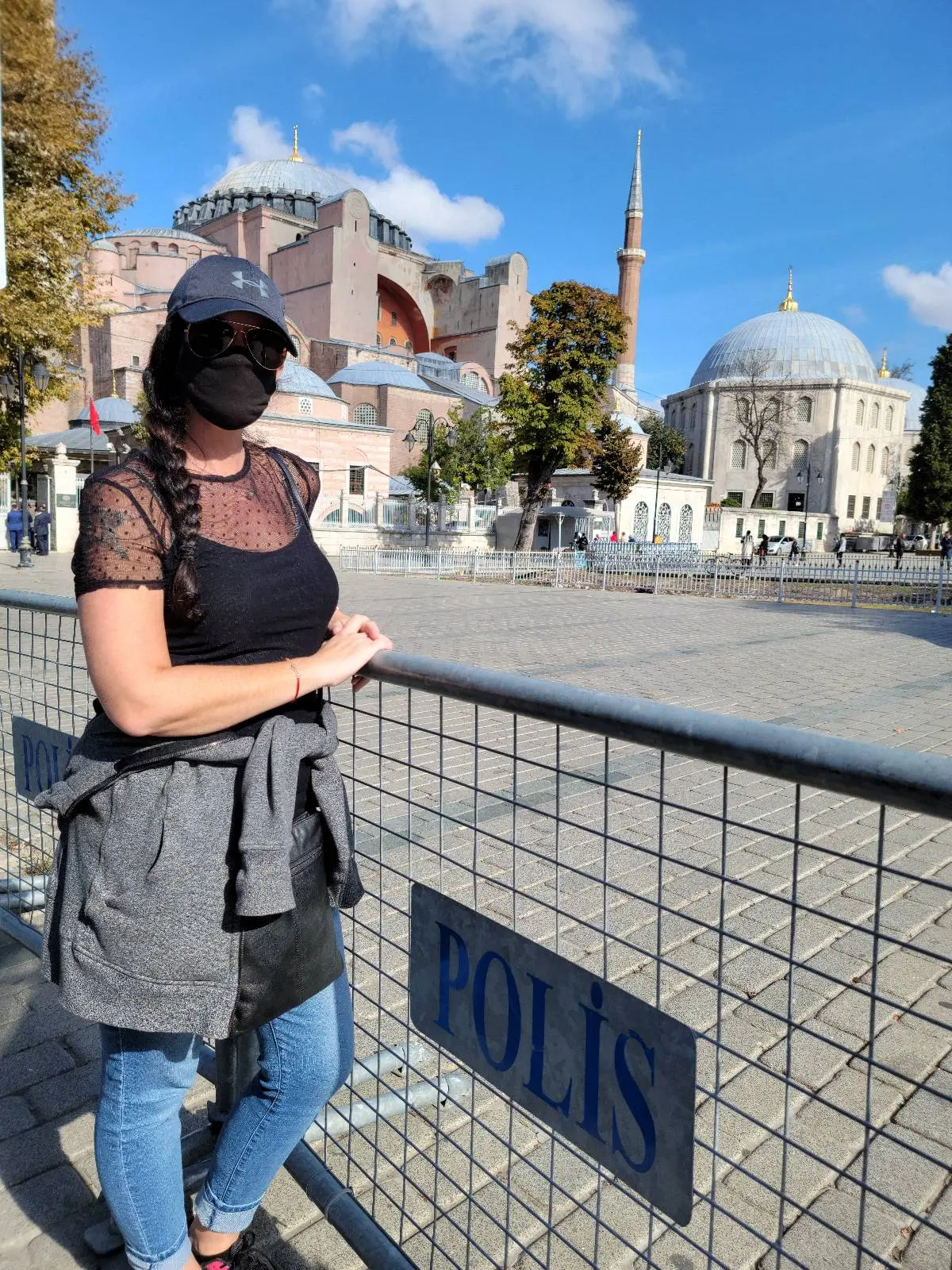 Person wearing a mask and cap stands by a fence with "Polis" sign, in front of a historic building with a dome and minarets.
