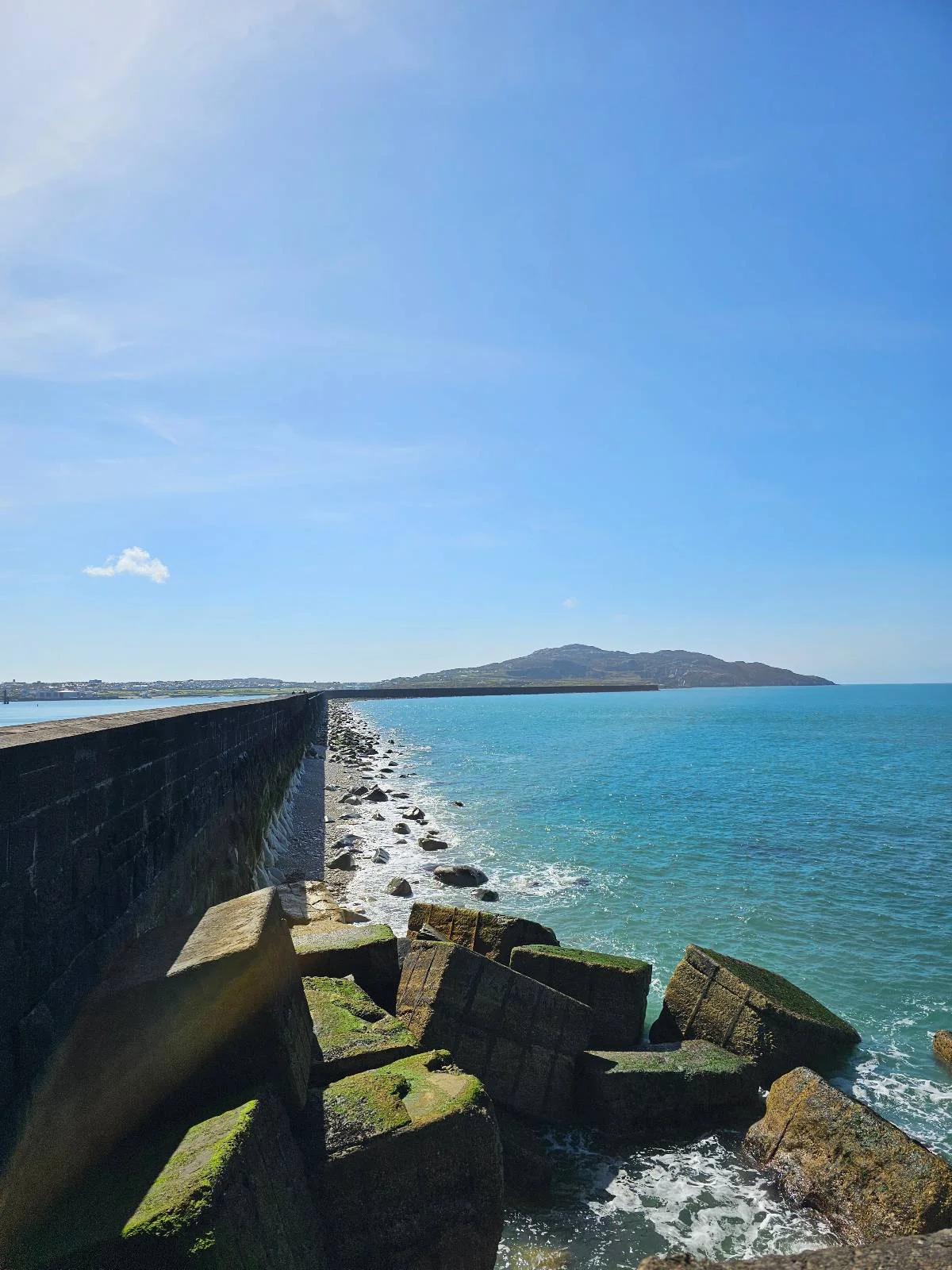 Coastal view showing a long seawall extending towards a distant mountain, under a clear blue sky with calm blue waters on the side.