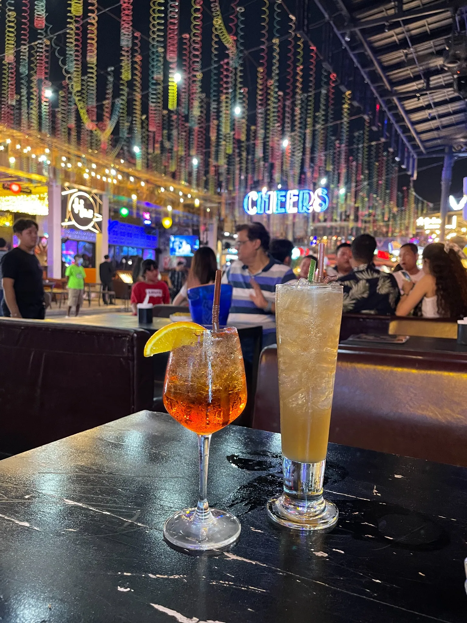 Two cocktails sit on a black marbled outdoor table at night; a tall iced drink with lime and a shorter orange drink. Neon city lights and blurred people are visible in the background.