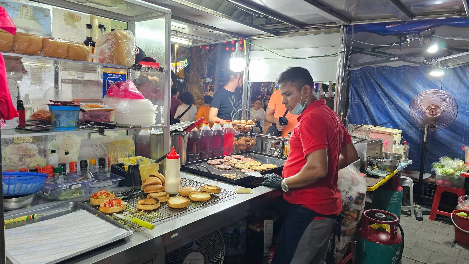 A street food vendor is wearing a red shirt and mask while grilling burgers at a food stall. The stall has shelves filled with ingredients, condiments, and buns. The setting is well-lit, and other stalls are visible in the background.