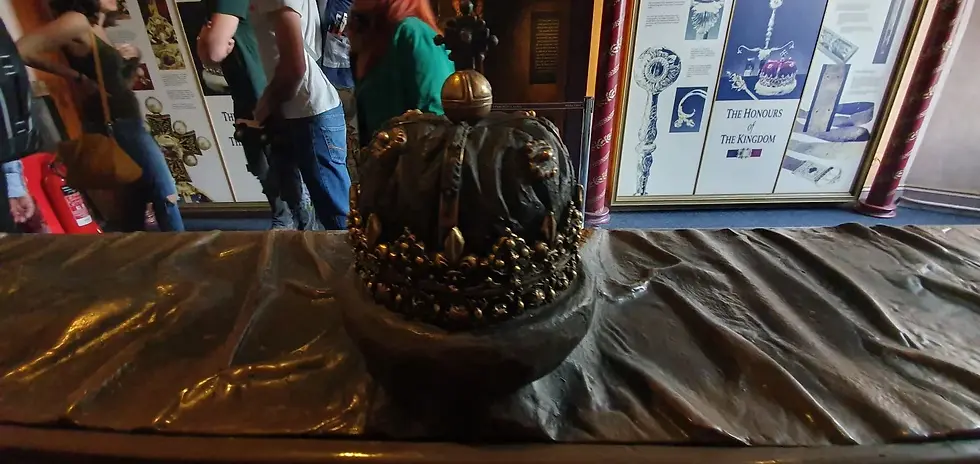 A gold crown with jewels is displayed on a draped table. People in the background view informational panels about royal honors.