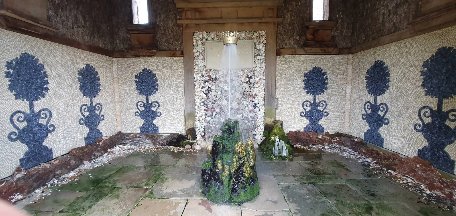 A decorative interior features mosaic walls with floral tree patterns in blue on a white background. The floor is made of stone tiles, with some greenery growing between them. A central stone structure stands in the middle of the room.