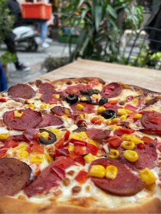 A close-up of a freshly baked pizza topped with pepperoni, black olives, red and yellow bell peppers, and melted cheese, with a blurred outdoor background.