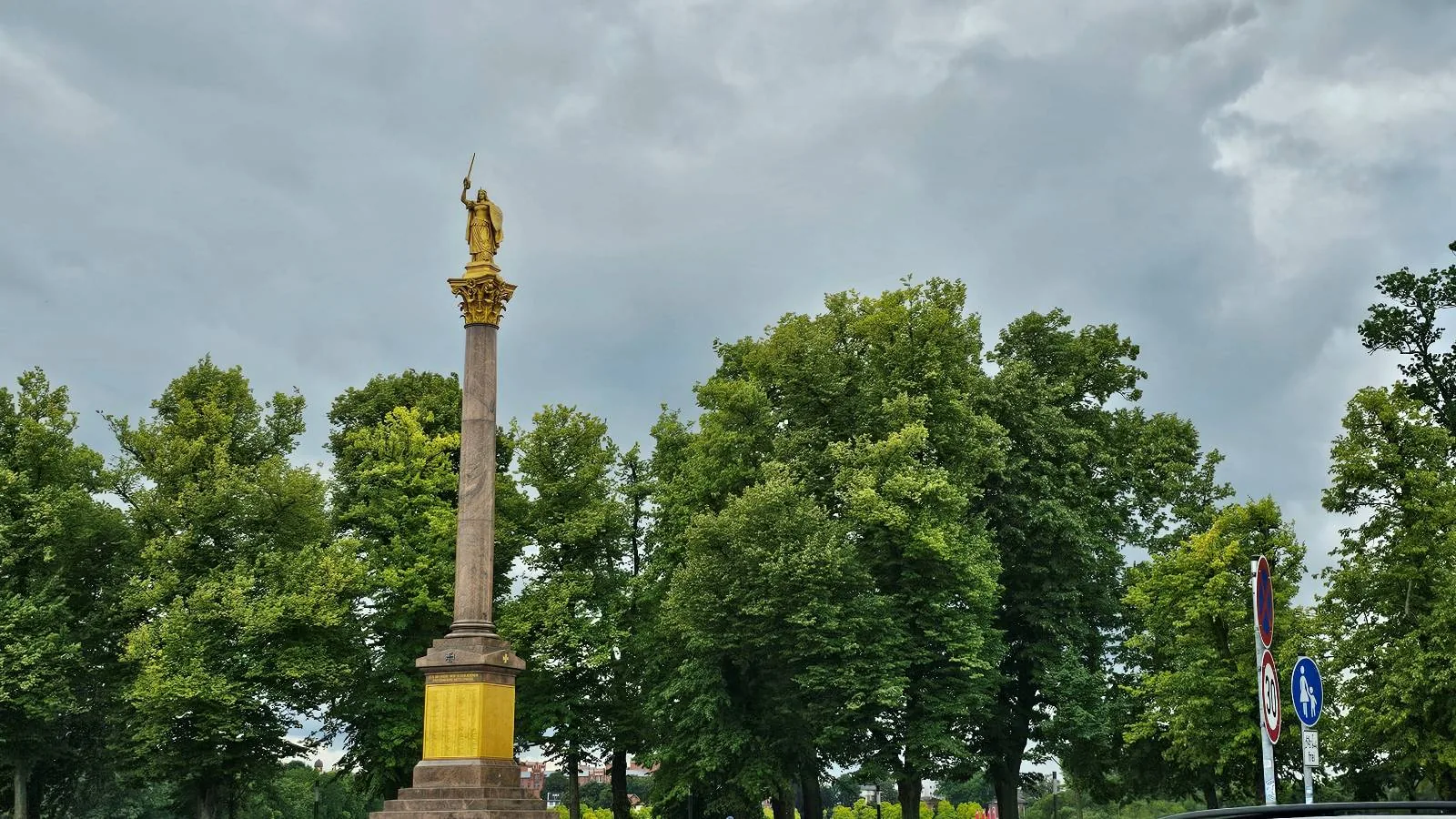 A tall column with a golden statue on top stands in front of lush green trees under a cloudy sky.
