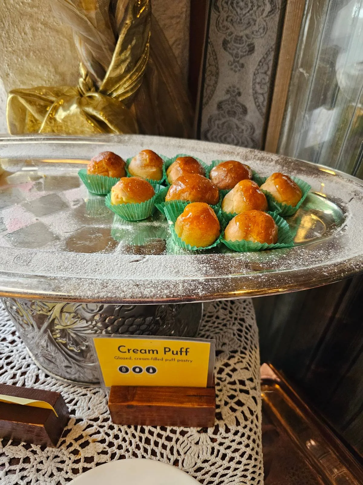 A tray of cream puffs in green wrappers on a decorative stand. A yellow sign with the label "Cream Puff" is displayed in front.