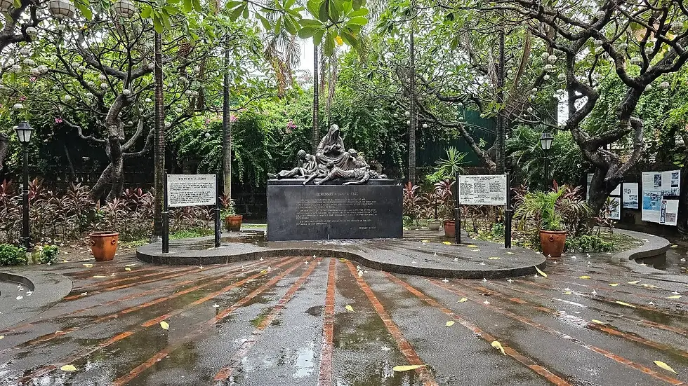 Bronze statue of a historic figure in a battle pose, surrounded by a wet circular paved area and lush green trees in an outdoor park setting.
