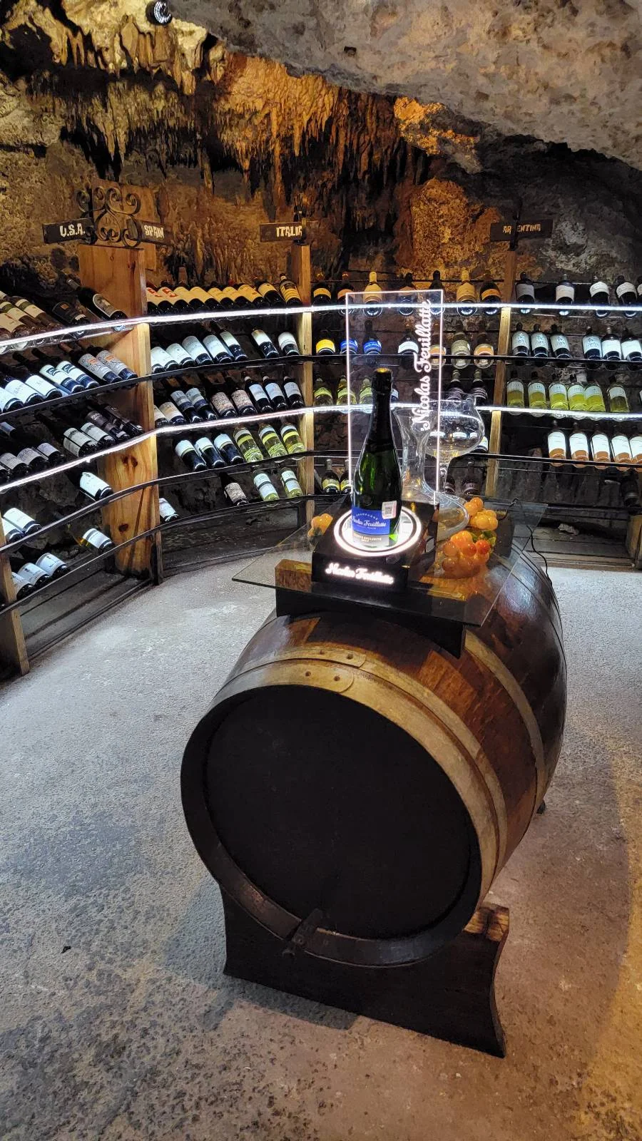 A wine cellar with bottles on wooden racks lining the walls. A large wooden barrel in the center holds a display with a standing wine bottle and two glasses. The ceiling has a rocky, cave-like appearance.