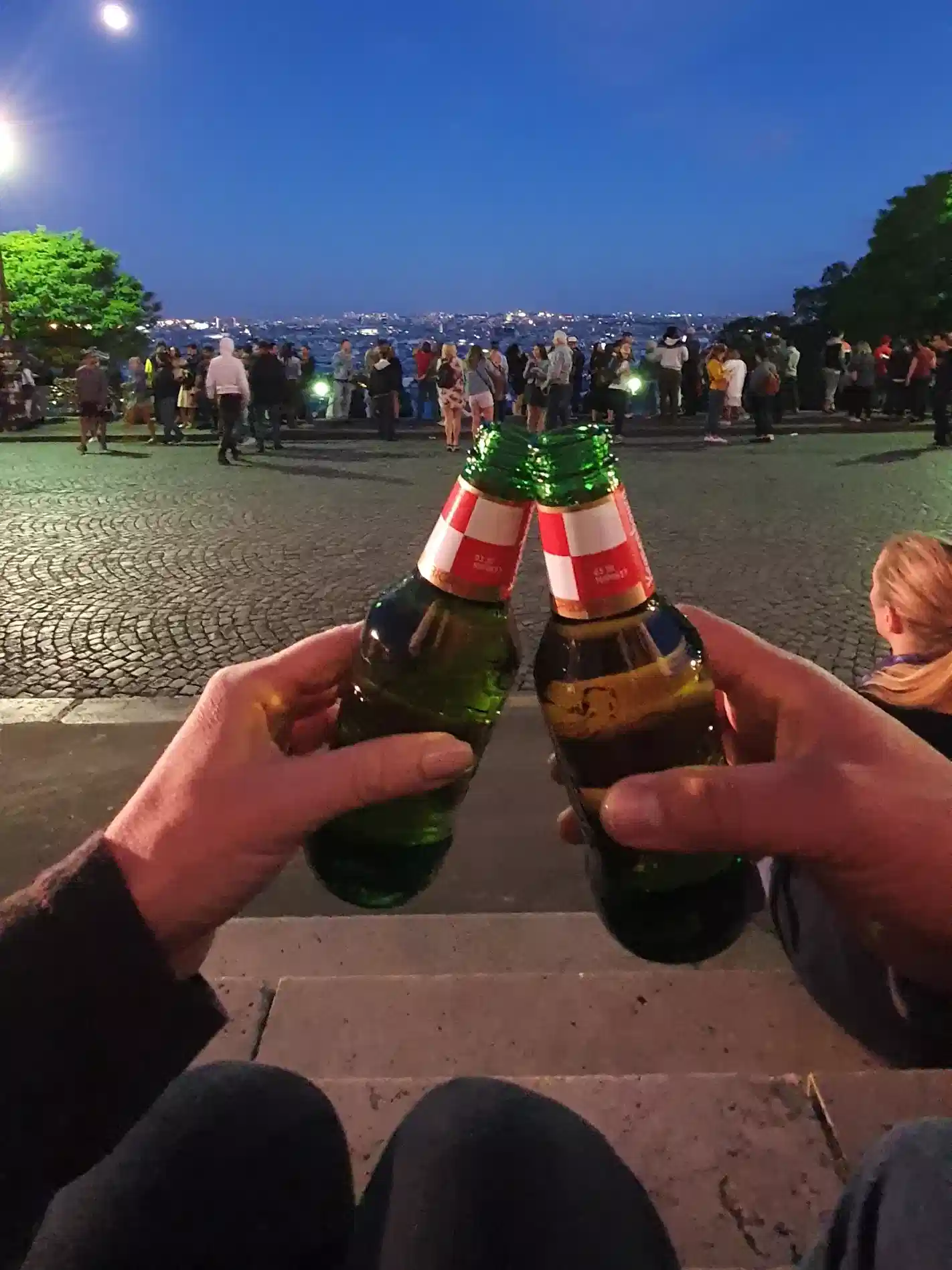 Two people clink green glass beer bottles together while sitting on steps at dusk, overlooking a street with a crowd of people and trees in the background.