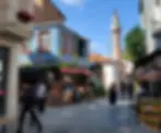 A bustling narrow street with shops, people walking, and a tall minaret in the background under a blue sky.